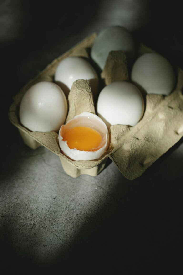 High angle of rustic carton container with uncooked fresh white whole and broken chicken eggs placed on table in sunlight